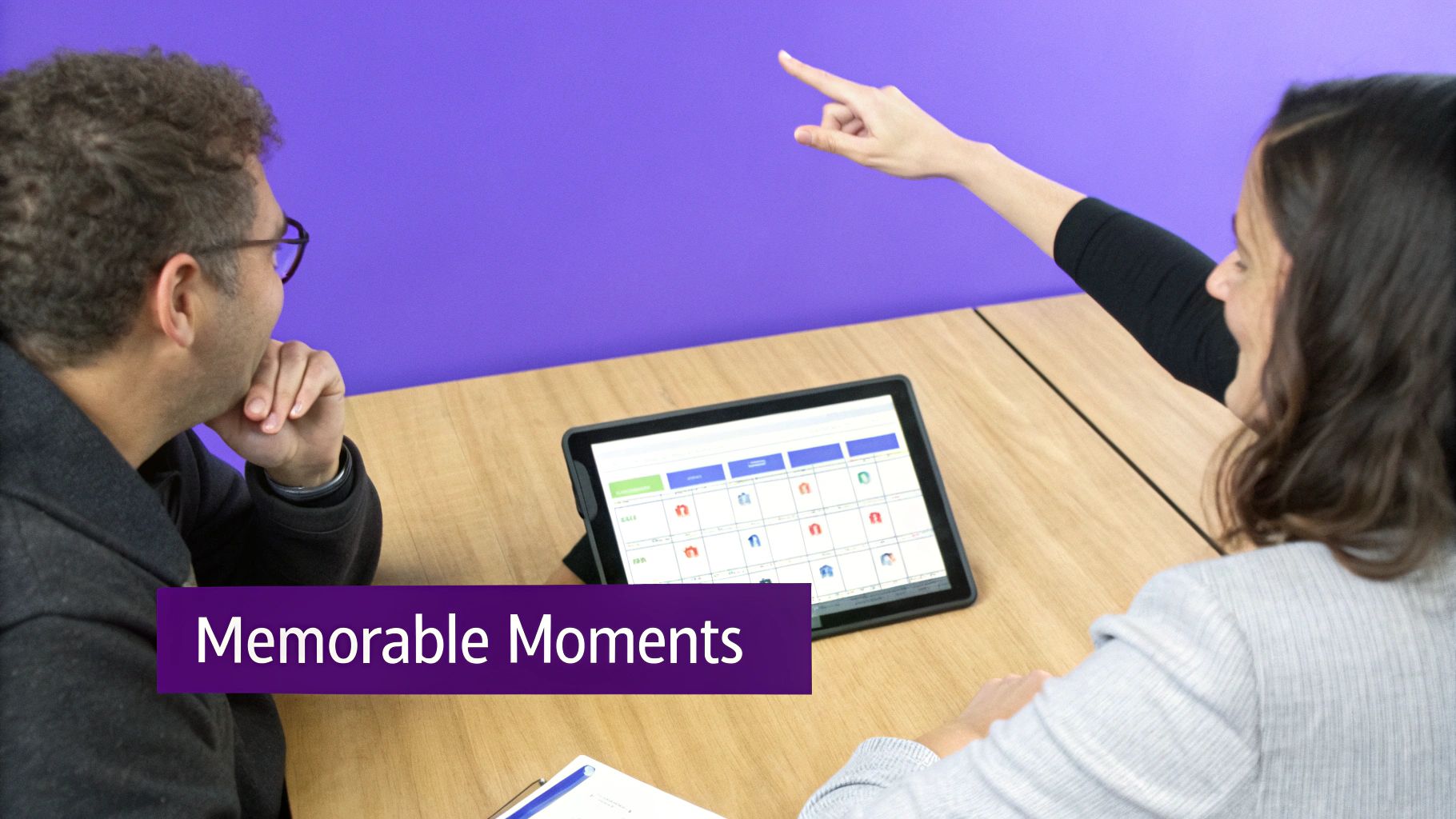 Two people discuss a digital schedule on a tablet at a table, with one person pointing.