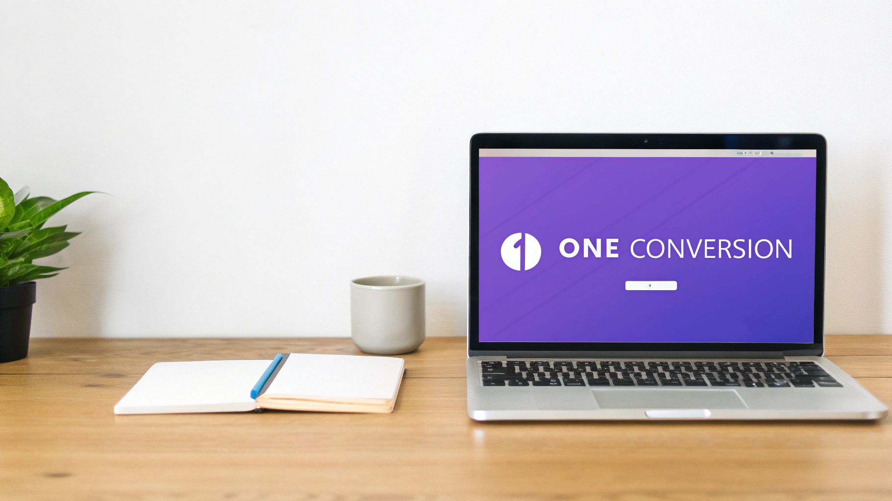 A minimalist workspace featuring a laptop displaying 'ONE CONVERSION', a plant, mug, and notebook on a wooden desk.
