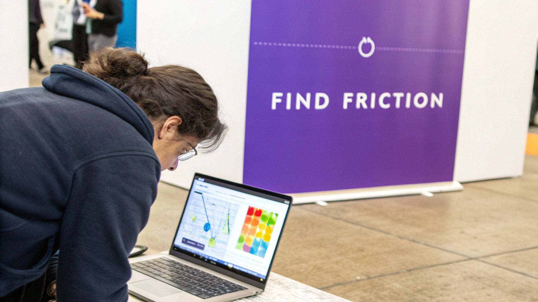 A person in a dark hoodie and glasses is focused on a laptop screen, with a purple banner in the background reading "FIND FRICTION".