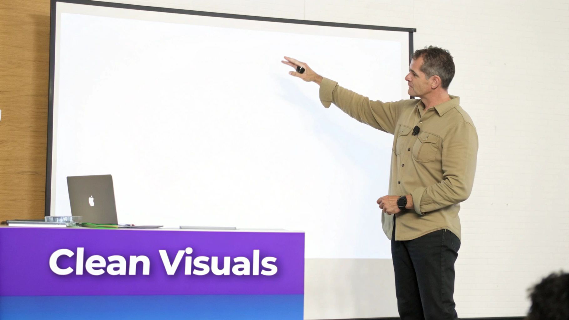 A man in a tan shirt presents in front of a blank screen, pointing, with a 'Clean Visuals' podium nearby.