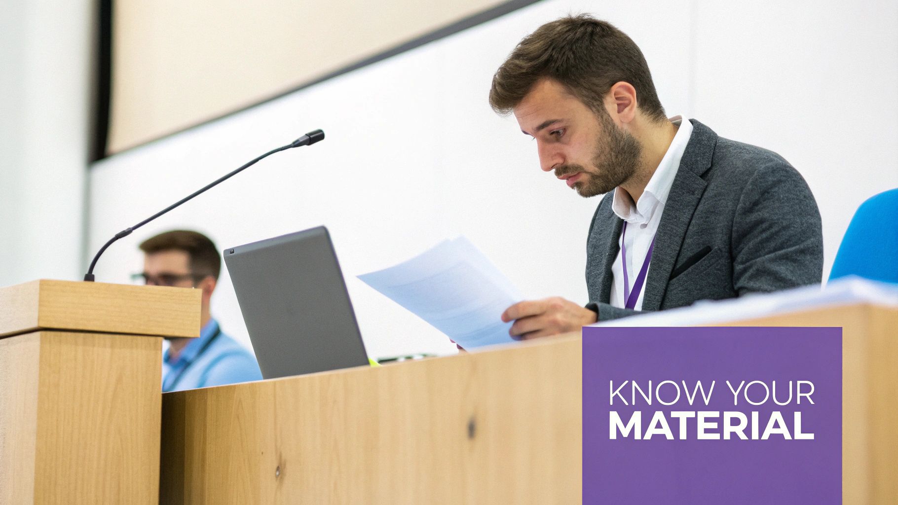 A man at a conference podium reviews documents with a laptop, focused on preparing his material.