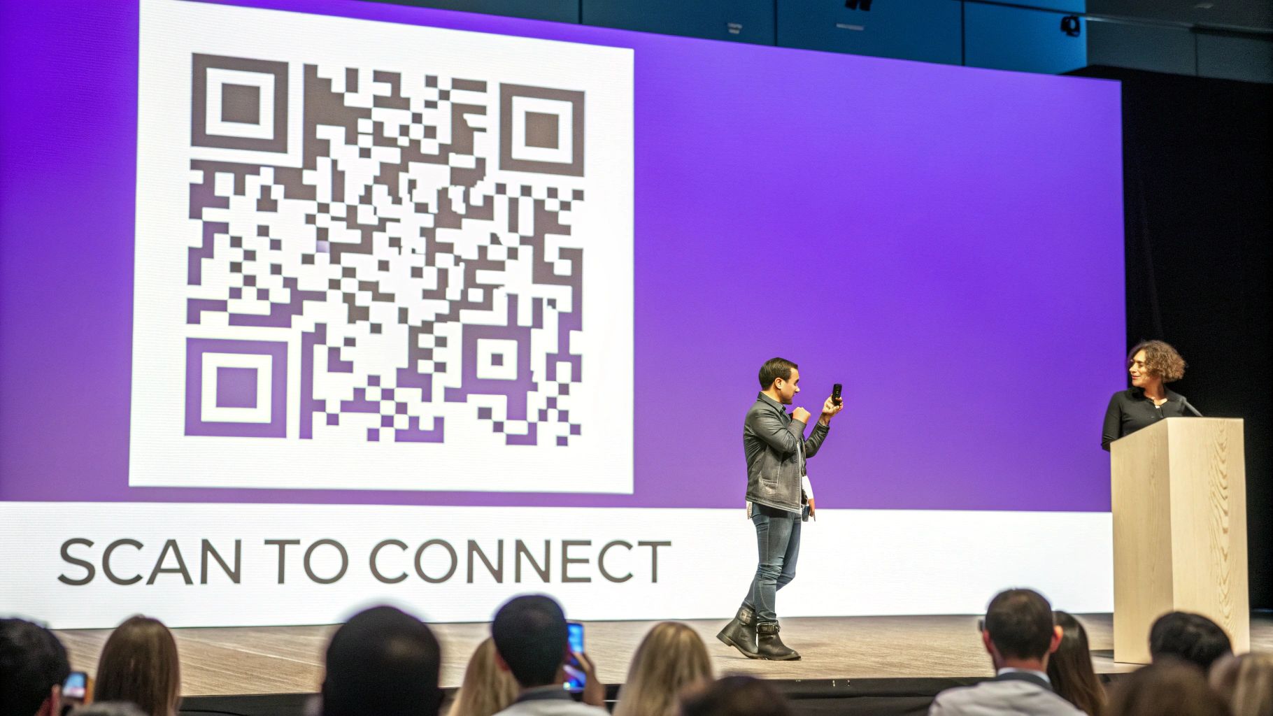 A speaker holds a phone towards a large screen displaying a QR code for audience connection.