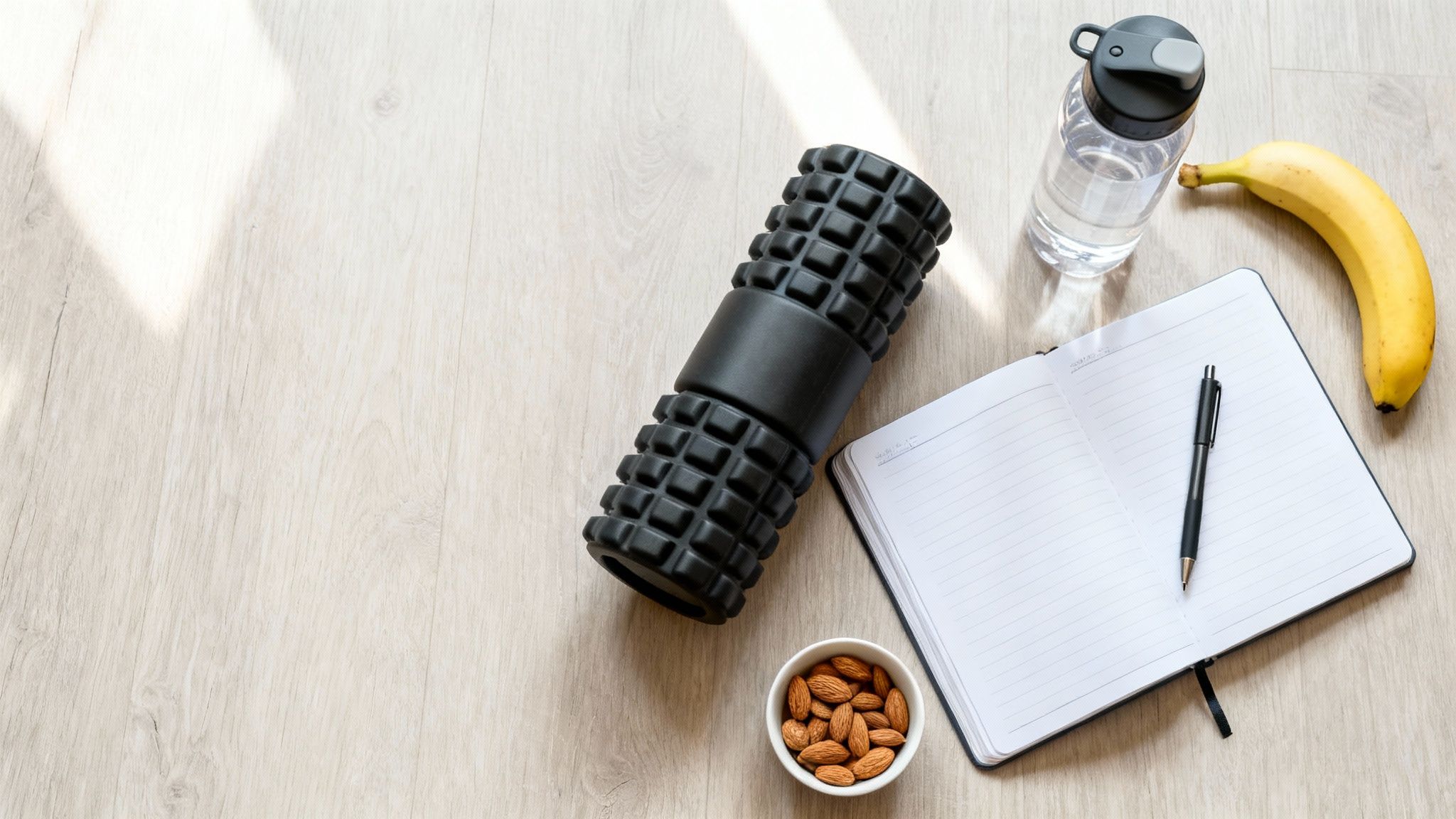 Flat lay of fitness gear and healthy snacks: foam roller, water, banana, almonds, and a notebook.