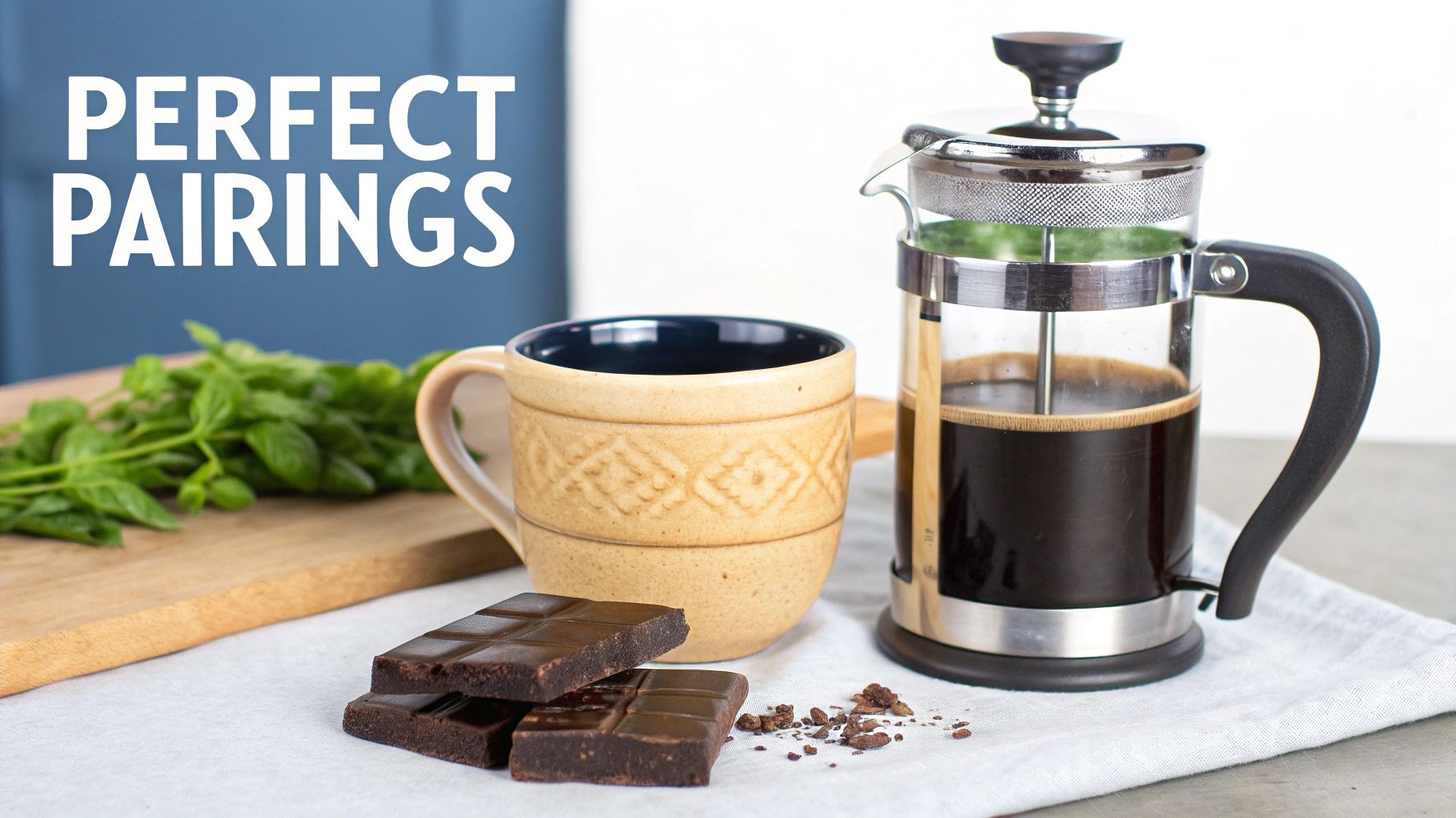A French press with coffee, a patterned mug, chocolate squares, and fresh mint leaves.