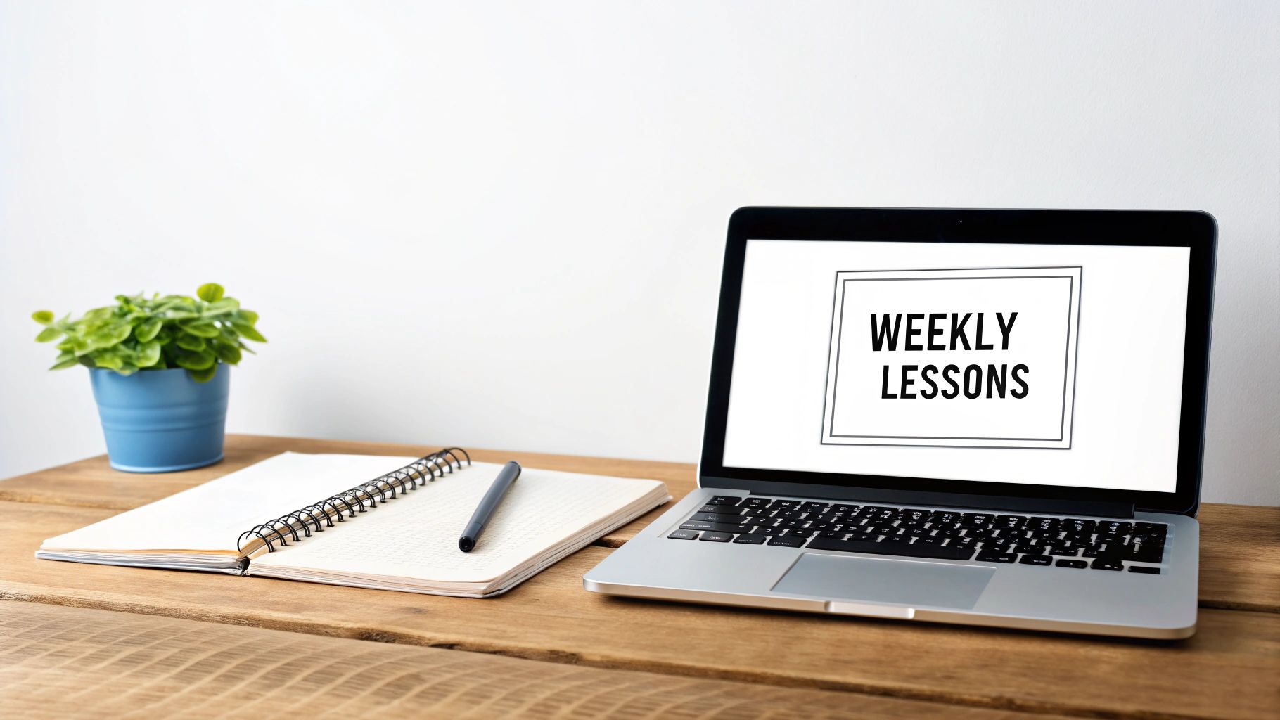 A laptop screen displays 'WEEKLY LESSONS' beside an open notebook, pen, and a potted plant on a wooden desk.