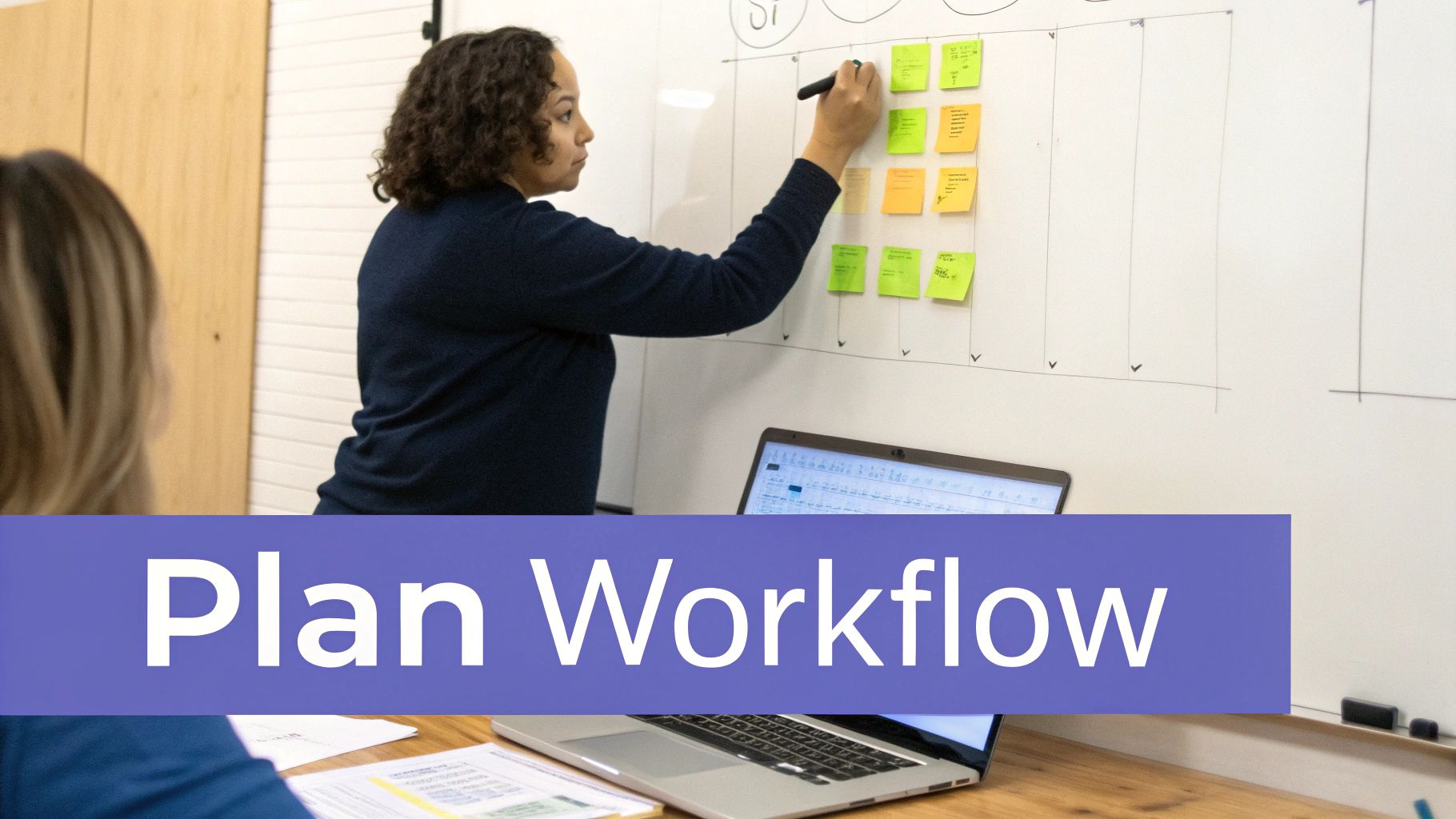 A woman plans a workflow on a whiteboard with sticky notes, while a colleague watches, with a laptop nearby.