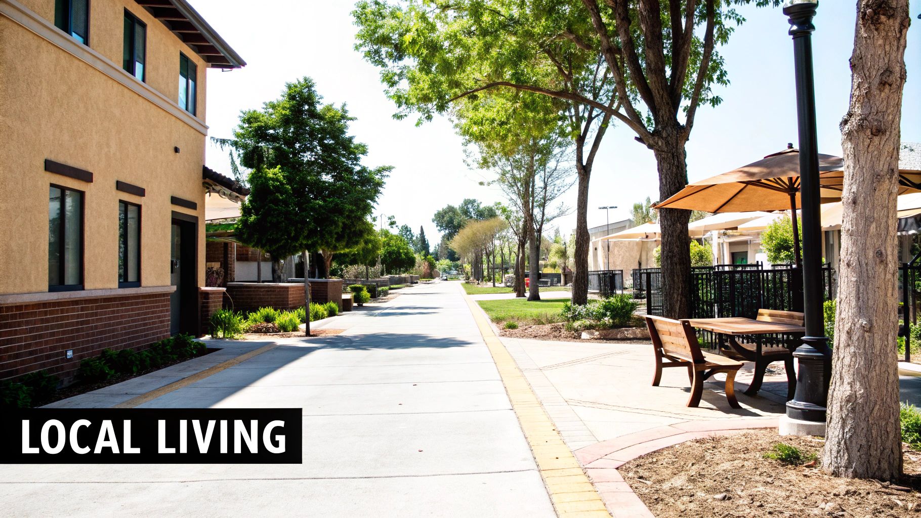 A sunny community street scene with a stucco building, tree-lined path, and outdoor patio with umbrella.
