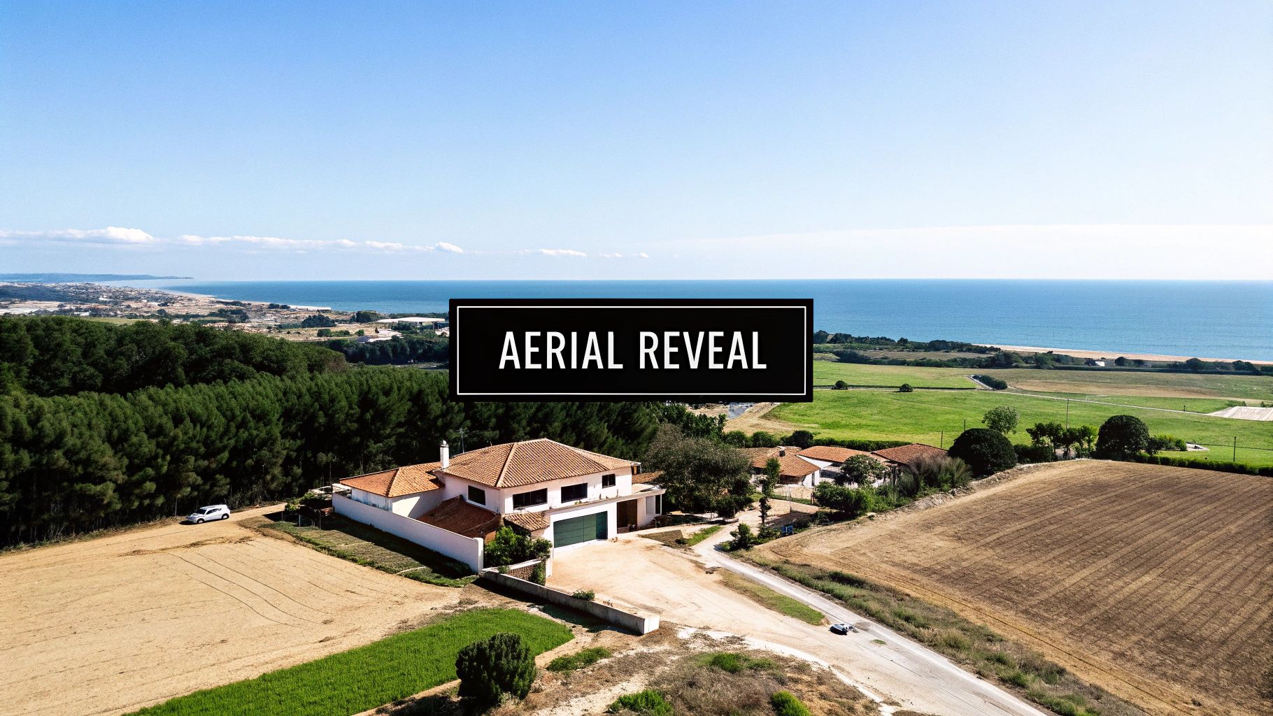Aerial view of a large house with a red-tiled roof, surrounded by fields, forest, and the ocean.