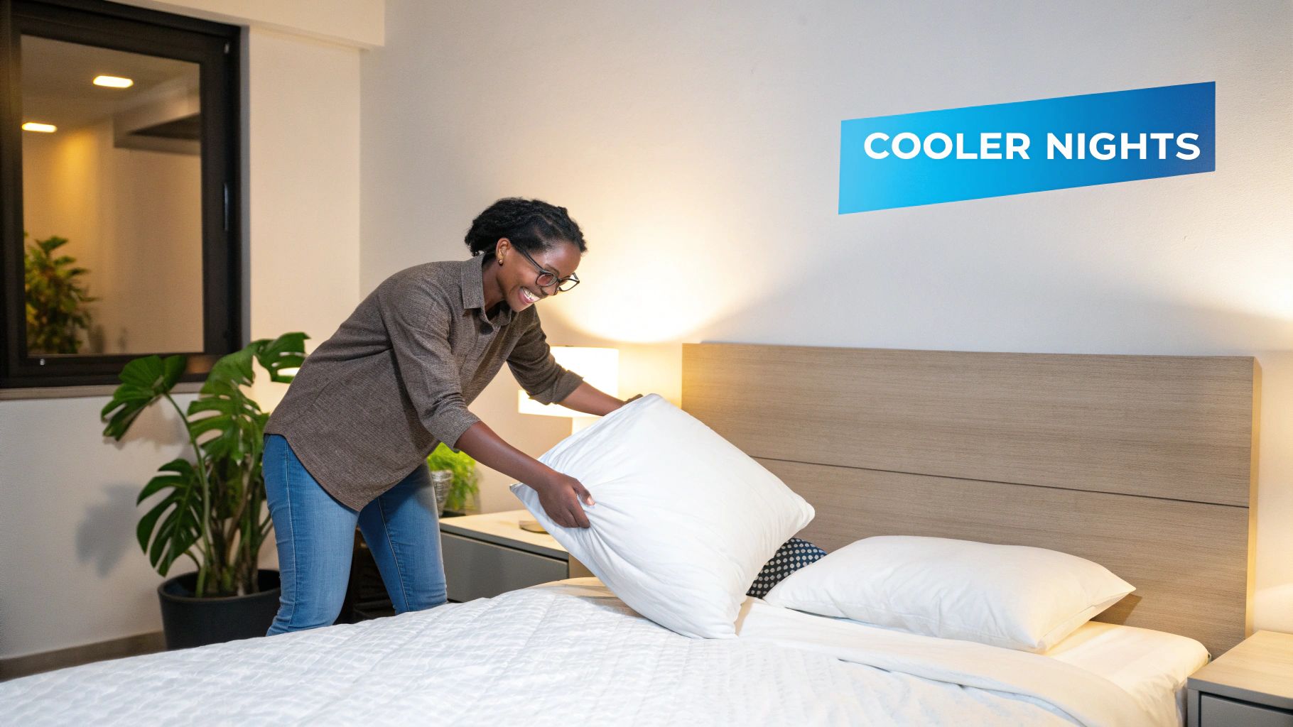 A happy woman in a modern bedroom arranges a white pillow on a freshly made bed.