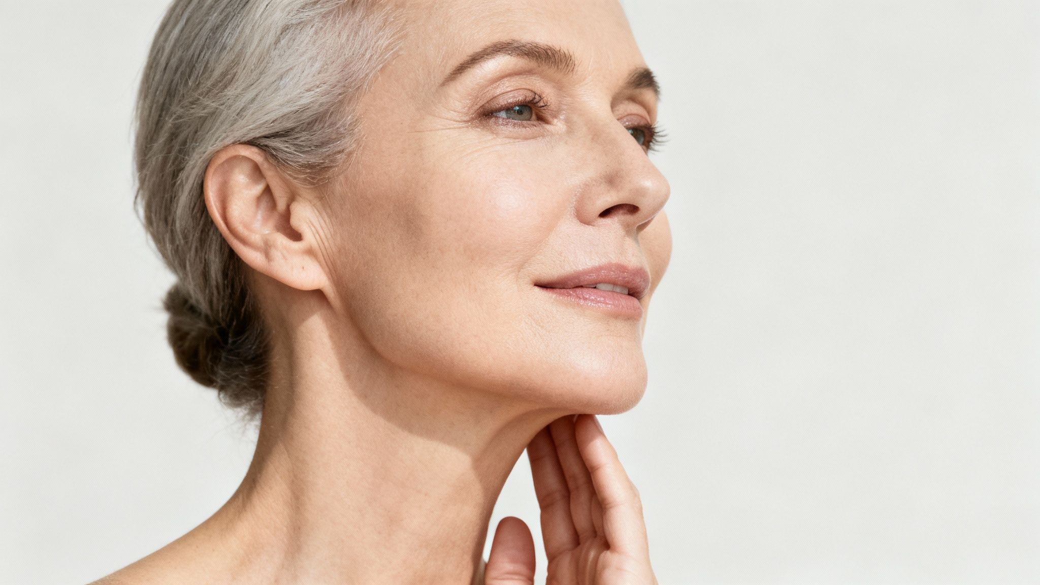 Side profile of an elegant mature woman with grey hair, touching her neck, with clear skin.