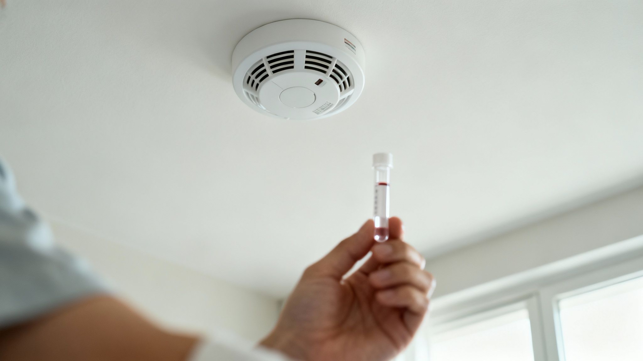 A hand holds a test tube with red liquid under a white smoke detector on a ceiling.