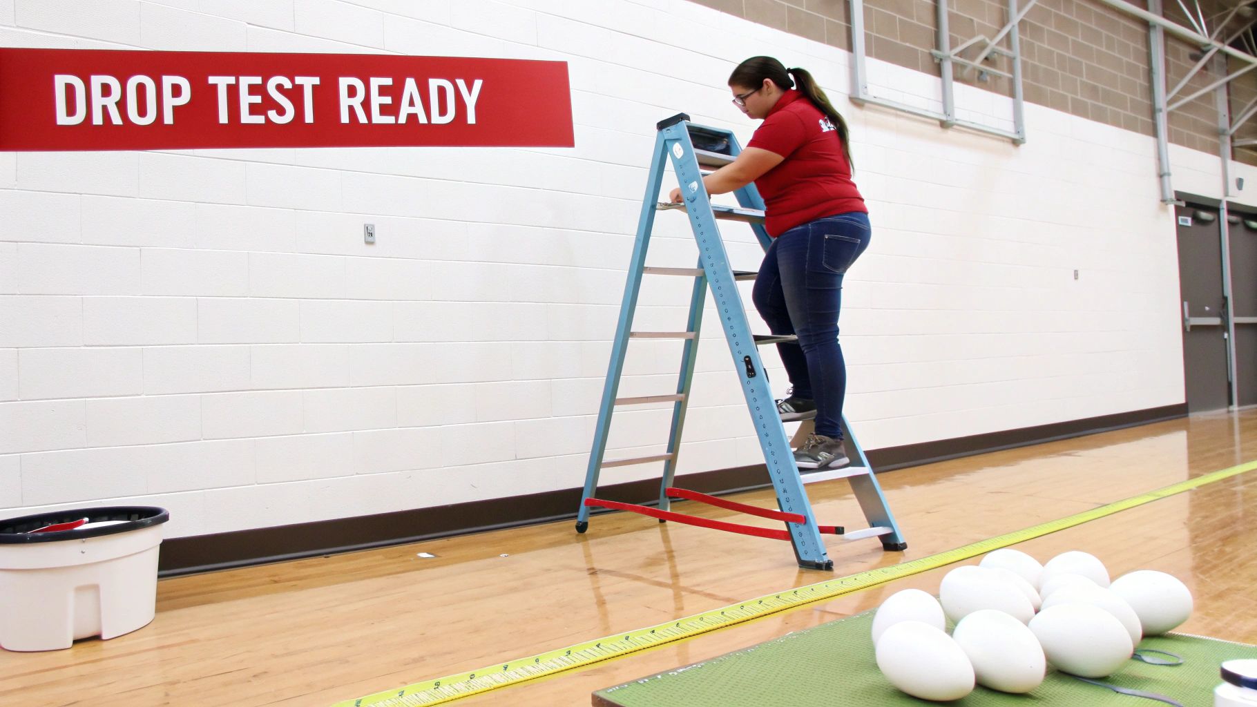 A student stands on a ladder next to a 'DROP TEST READY' banner, preparing for an egg experiment.