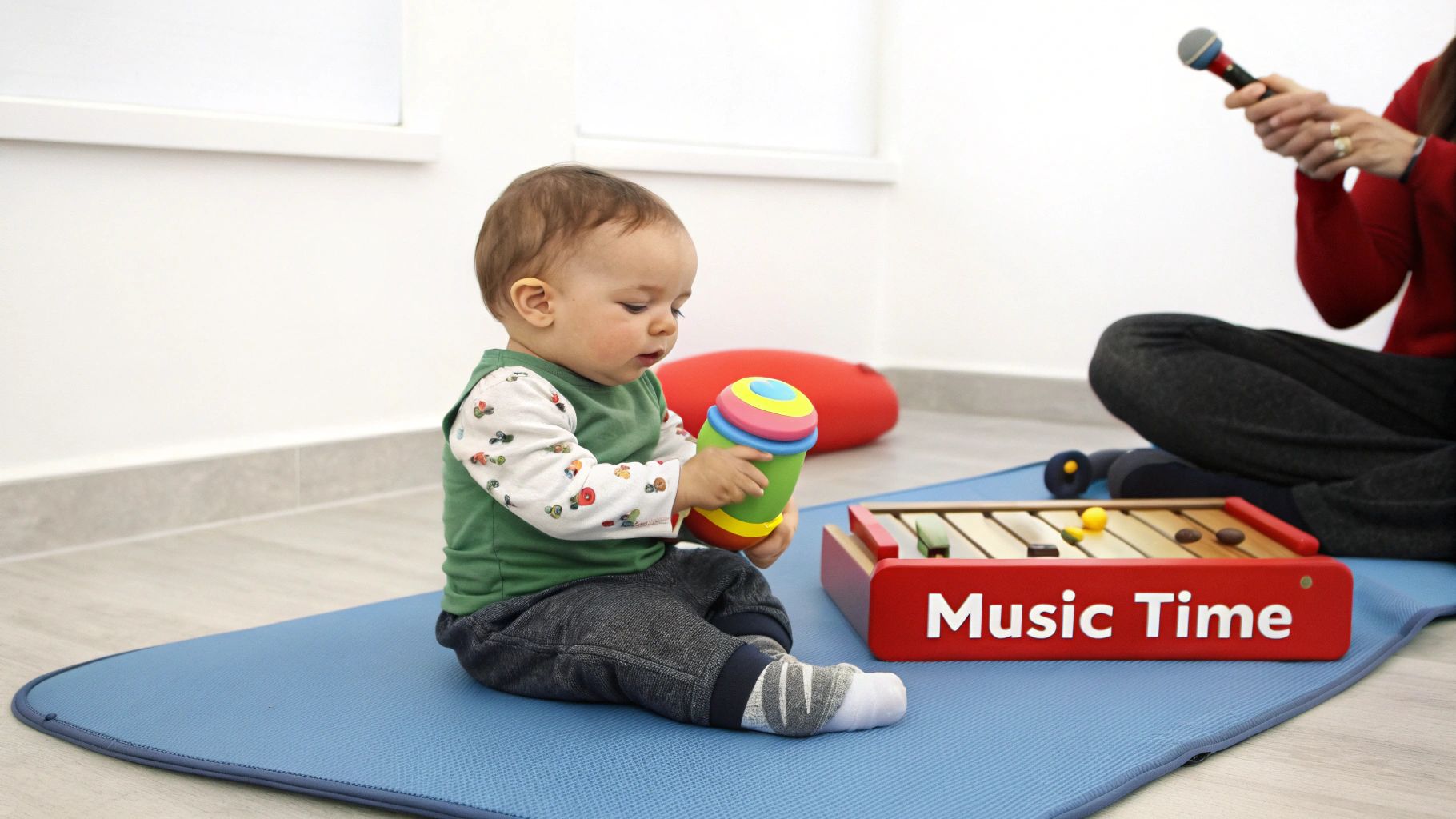 A baby sits on a blue mat, playing with a colorful rattle during music time with an adult.