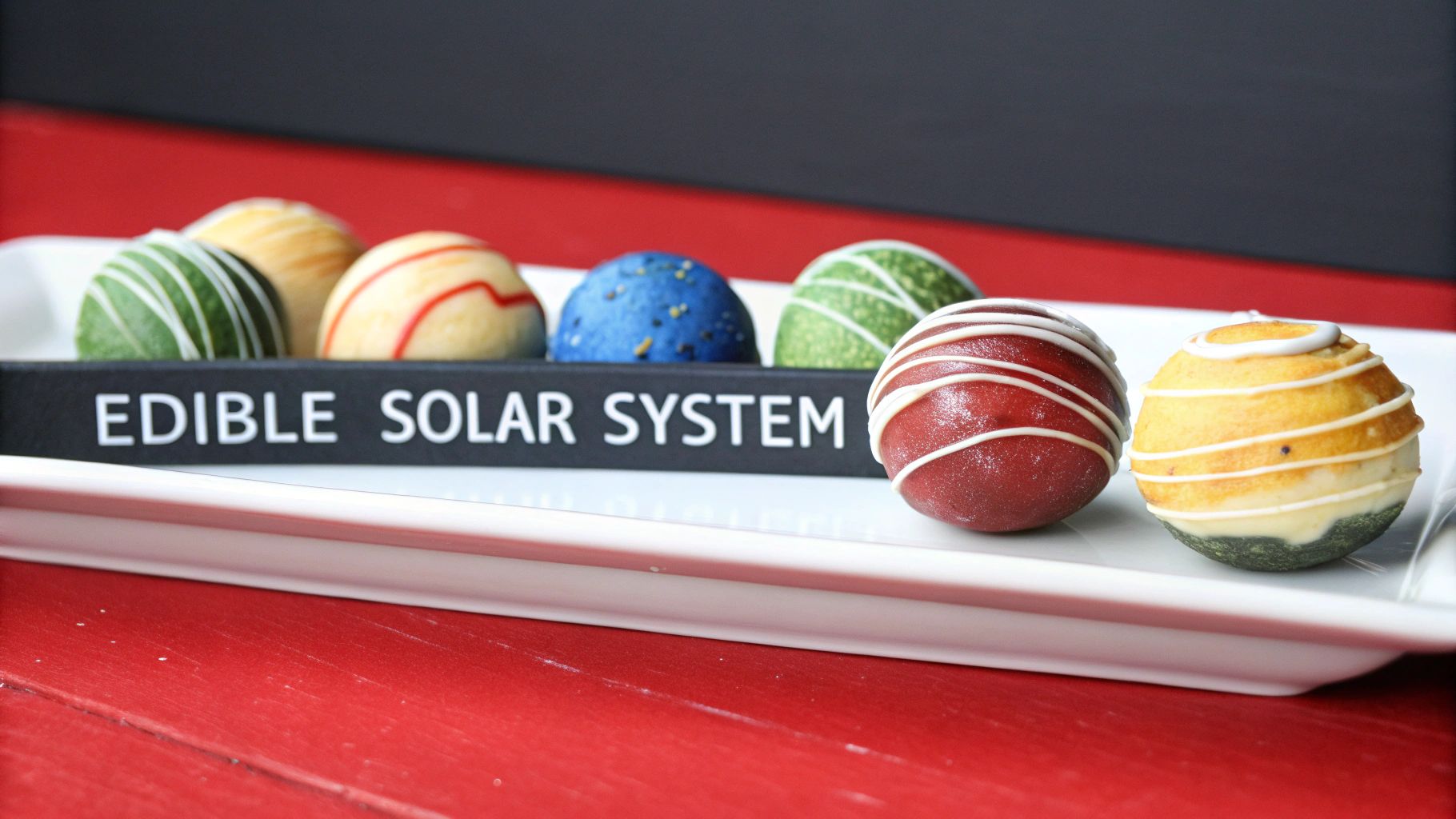 A line of colorful, decorated edible treats, resembling planets, on a white tray labeled "Edible Solar System."