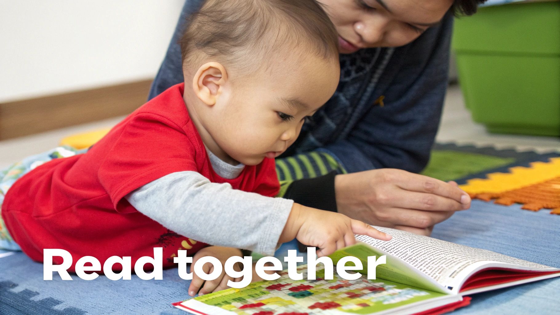 A baby in a red shirt and an adult read a colorful book together on the floor.