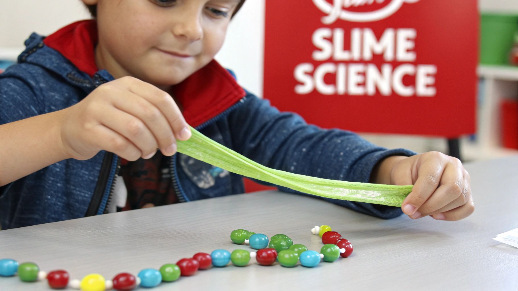 A child happily stretches green slime at a science activity table with colorful beads.