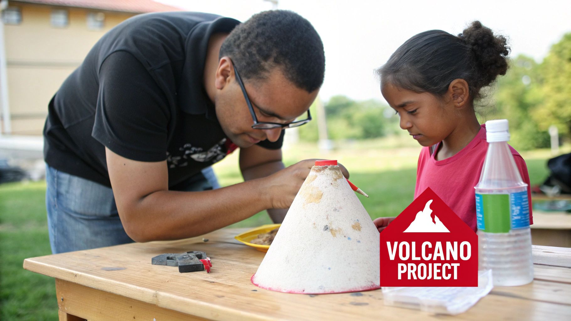 An adult helps a young girl build a volcano model on a wooden table outdoors.