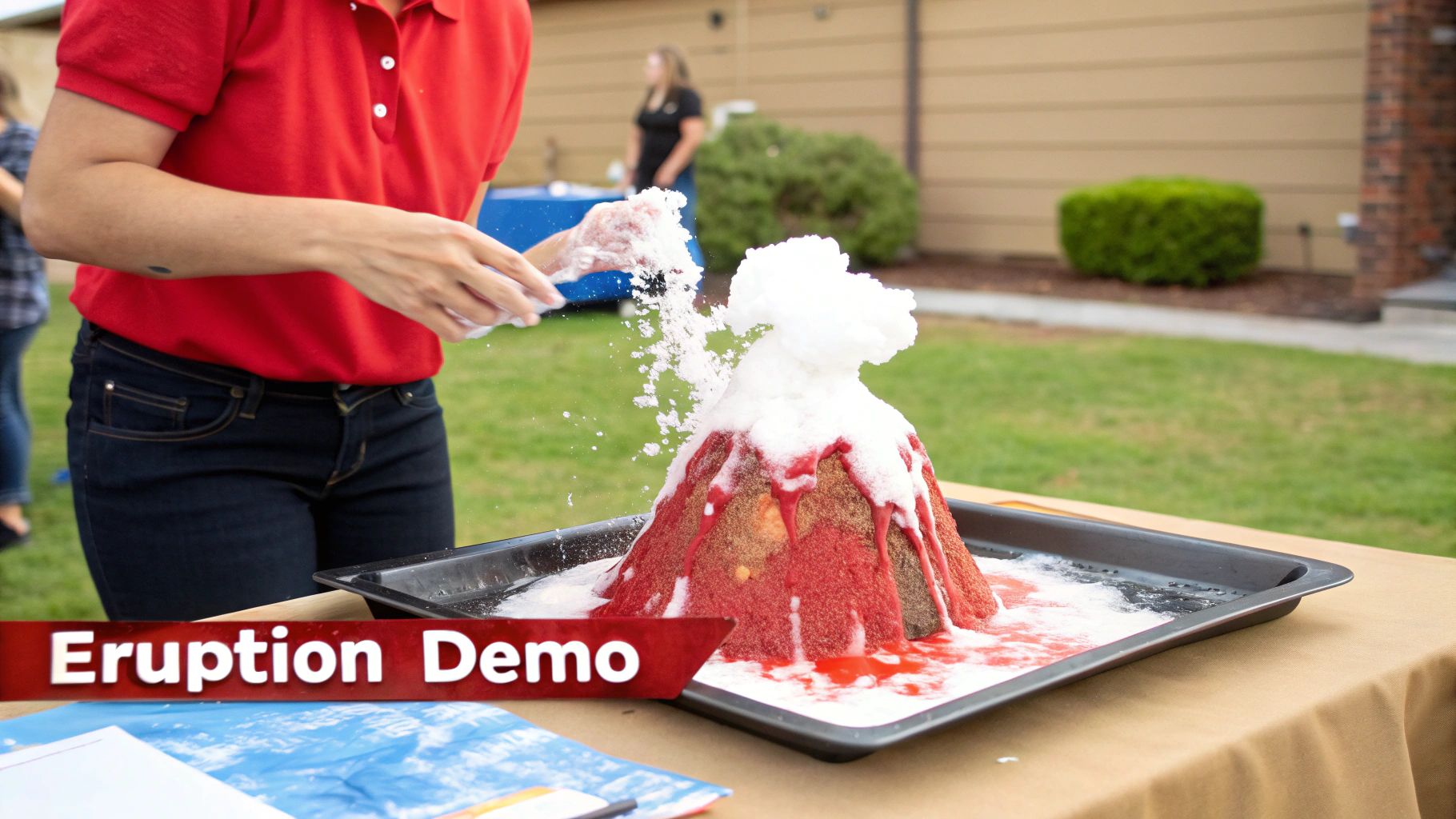 A person in a red shirt demonstrates a volcano model erupting with foam and red liquid outdoors.