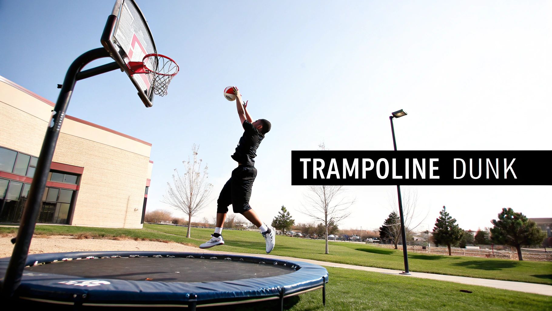 A person in black sportswear jumps high on a trampoline, attempting to dunk a basketball into an outdoor hoop.