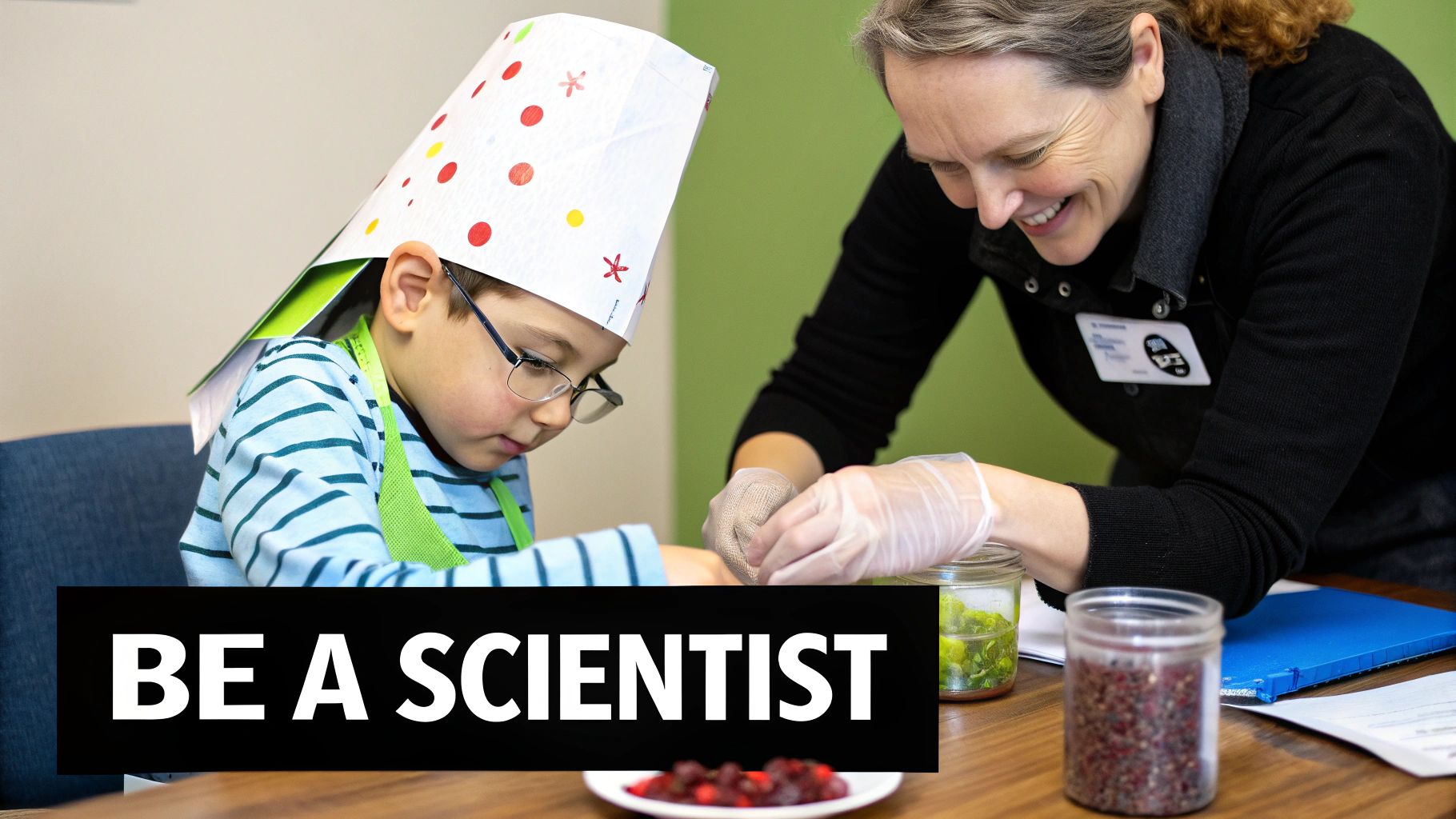 A young boy with glasses and a chef's hat, assisted by a smiling adult, performs a science activity.