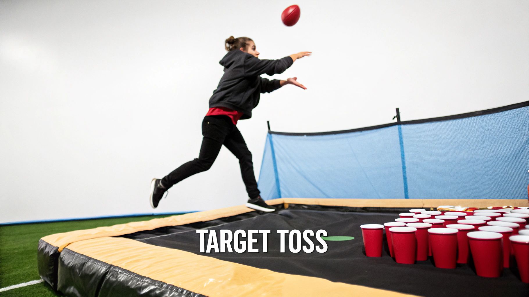 A kid leaps, throwing a red football at red plastic cups on a trampoline game.