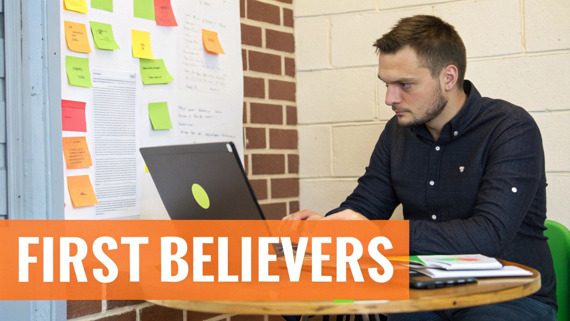 Focused man in a dark shirt working on a laptop at a table, with a whiteboard full of sticky notes behind him.