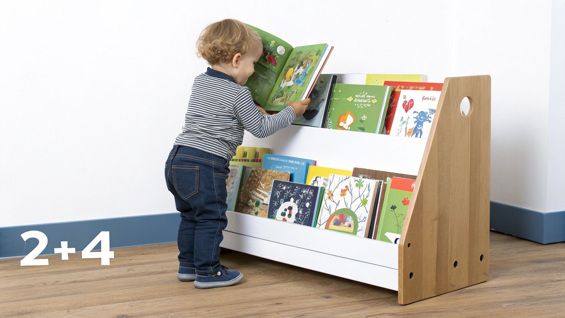 A toddler in a striped shirt and jeans reads a book on a white and wooden children's bookcase.
