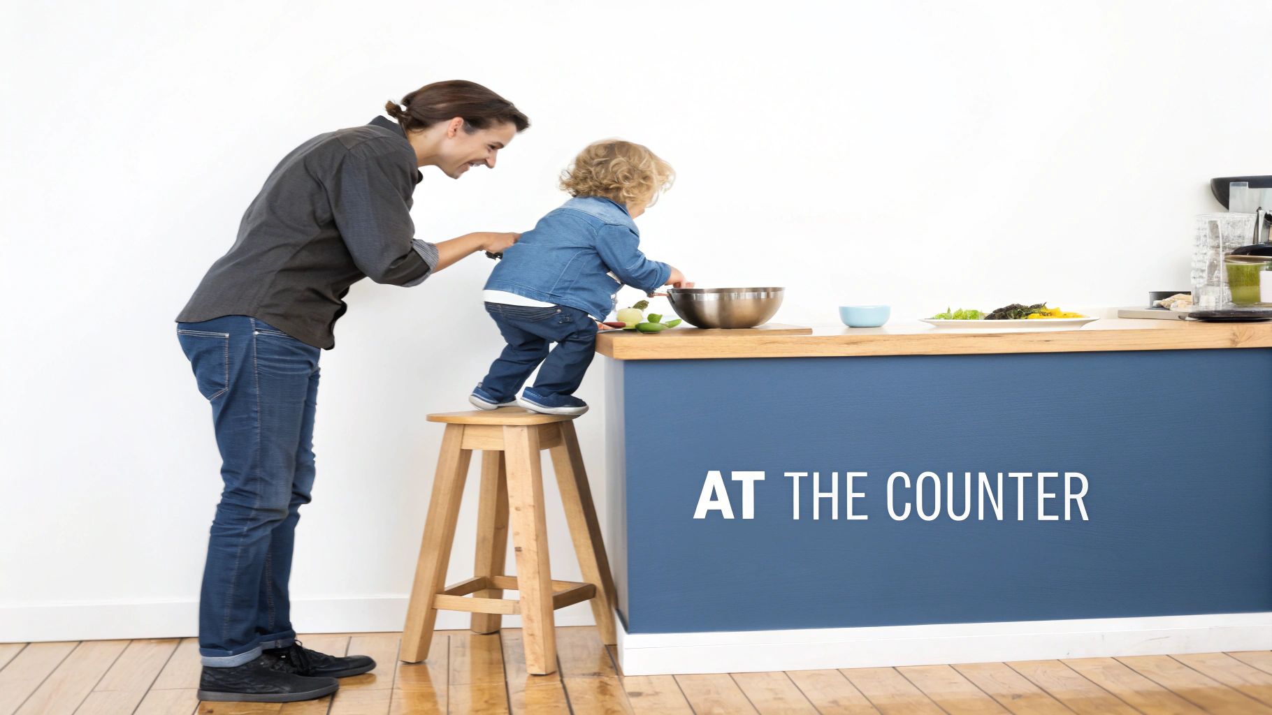 Smiling adult assists a toddler learning to cook on a stool at a modern kitchen counter.