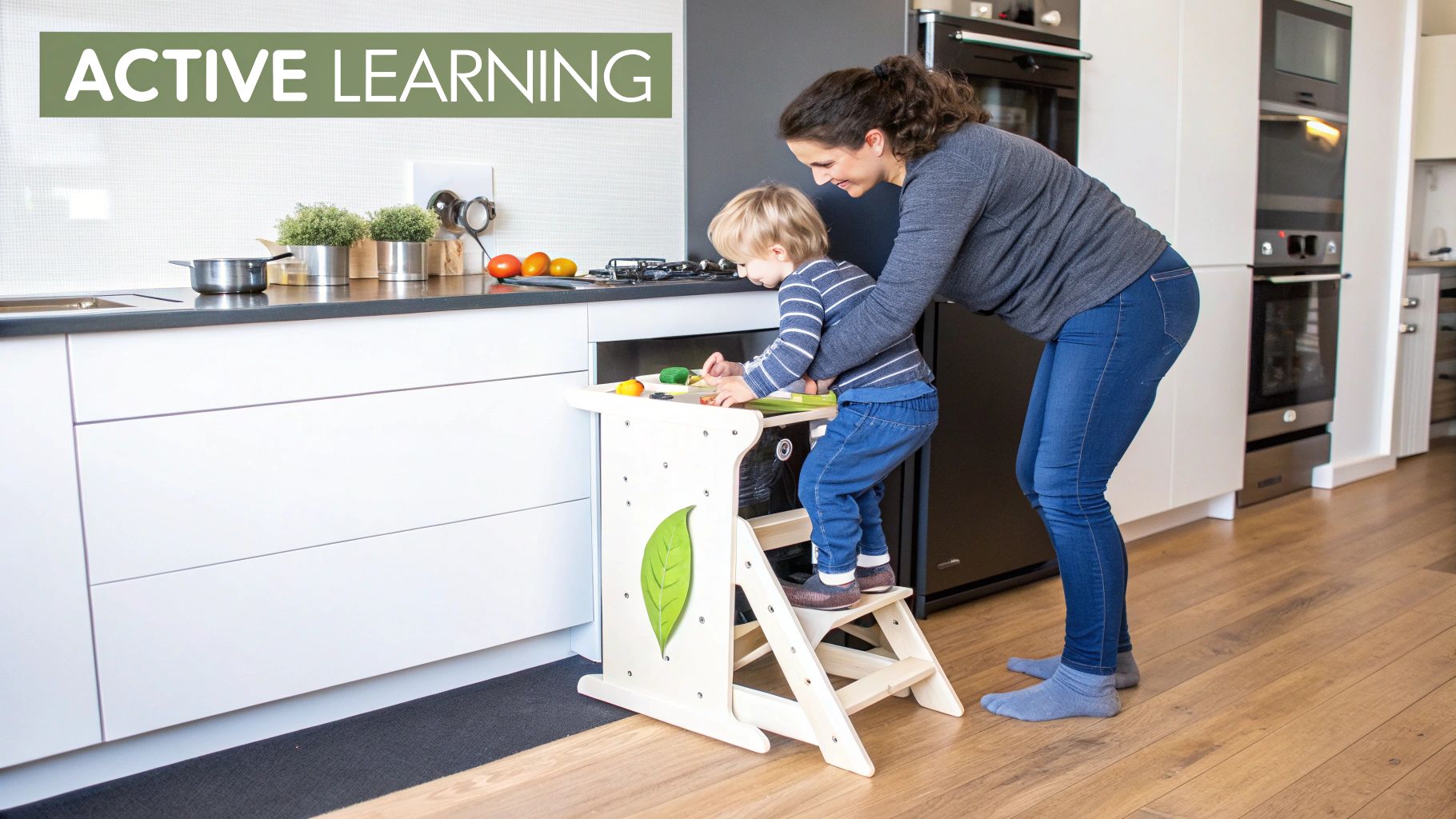 A young child stands in a wooden learning tower, smiling and helping prepare food at a kitchen counter.