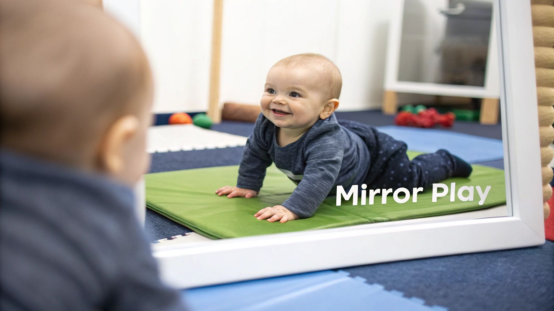 A smiling baby on a green mat looks at its reflection in a mirror during tummy time.