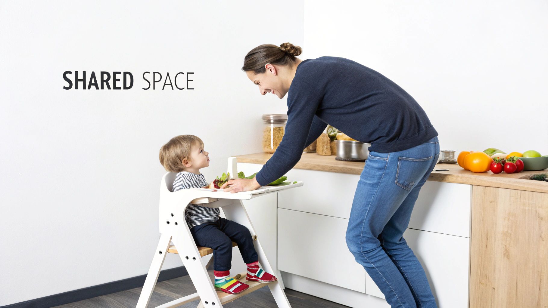 A toddler stands in a wooden learning tower at a kitchen counter, helping a parent prepare food.