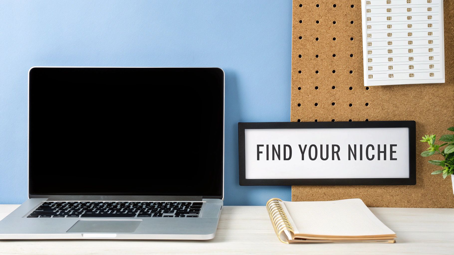 A desk setup featuring a laptop, an open notebook, and a 'FIND YOUR NICHE' sign.