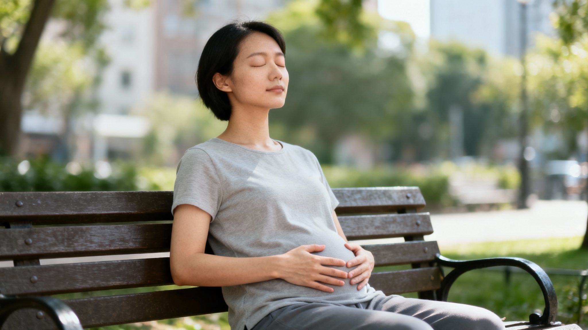 Pregnant Asian woman meditating peacefully on a park bench, hands on her belly.