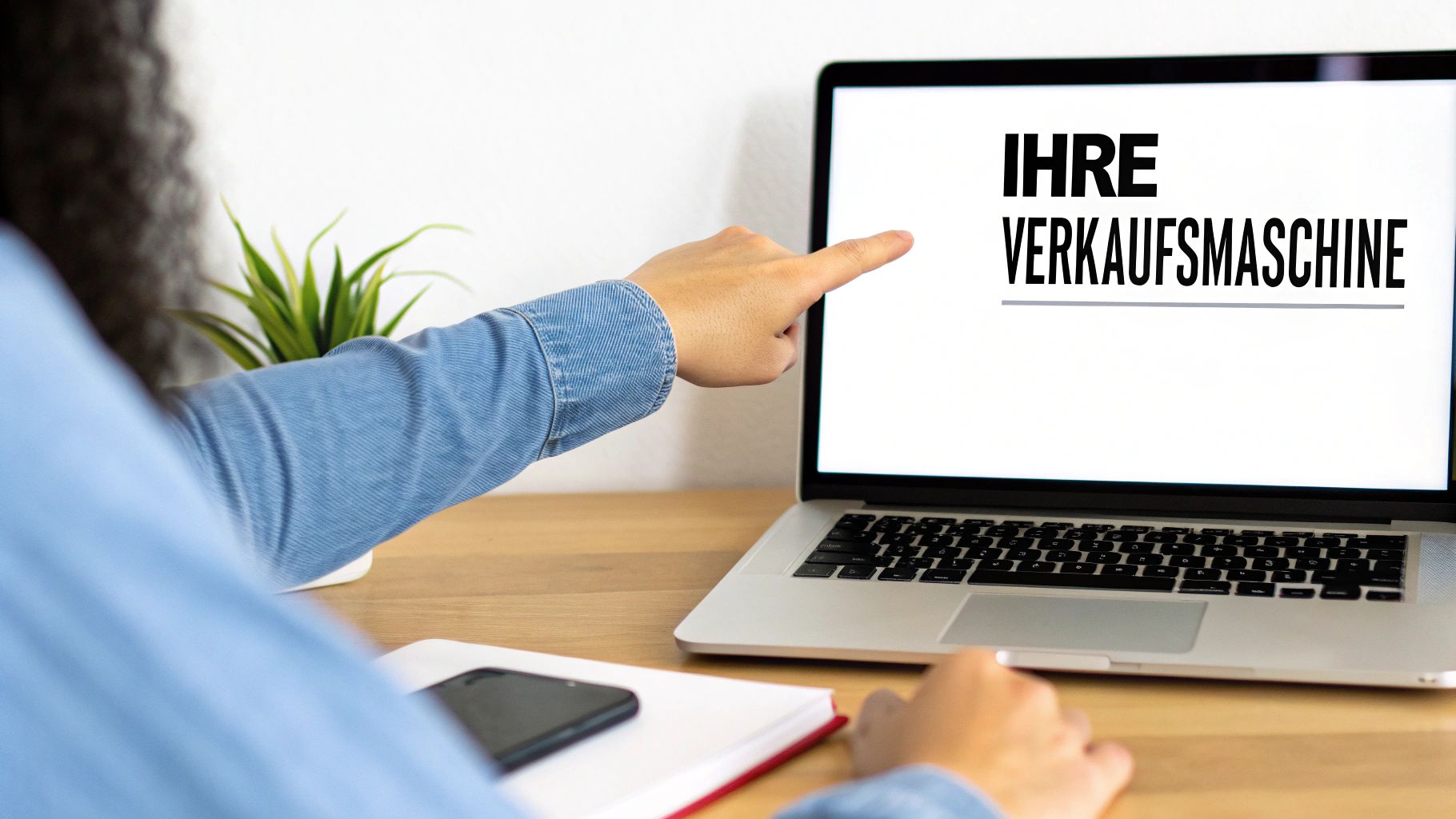 Eine Person zeigt auf einen Laptop mit dem Text 'IHRE VERKAUFSMASCHINE' auf dem Bildschirm, auf einem Holzschreibtisch.