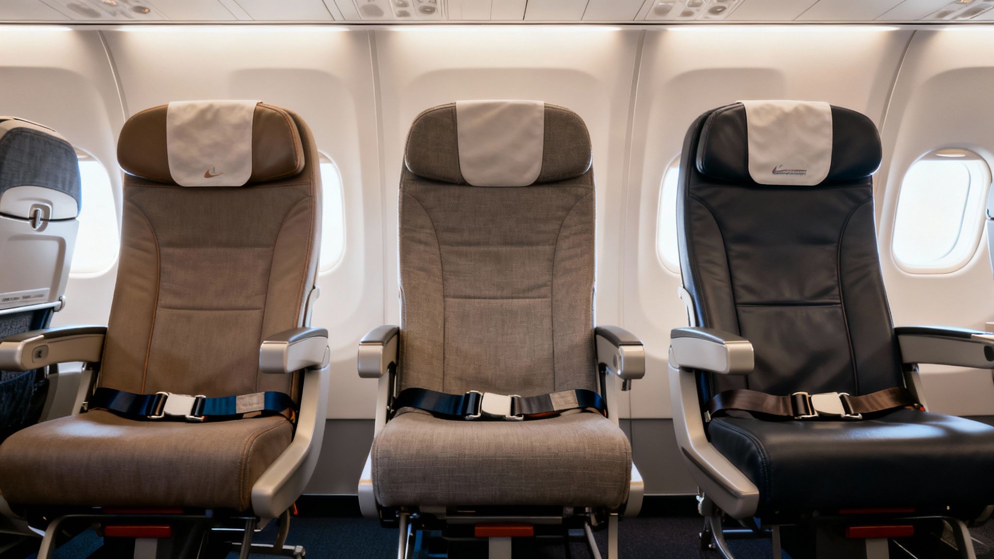 Three empty airplane seats with visible seatbelts, showing different upholstery colors and headrest covers.