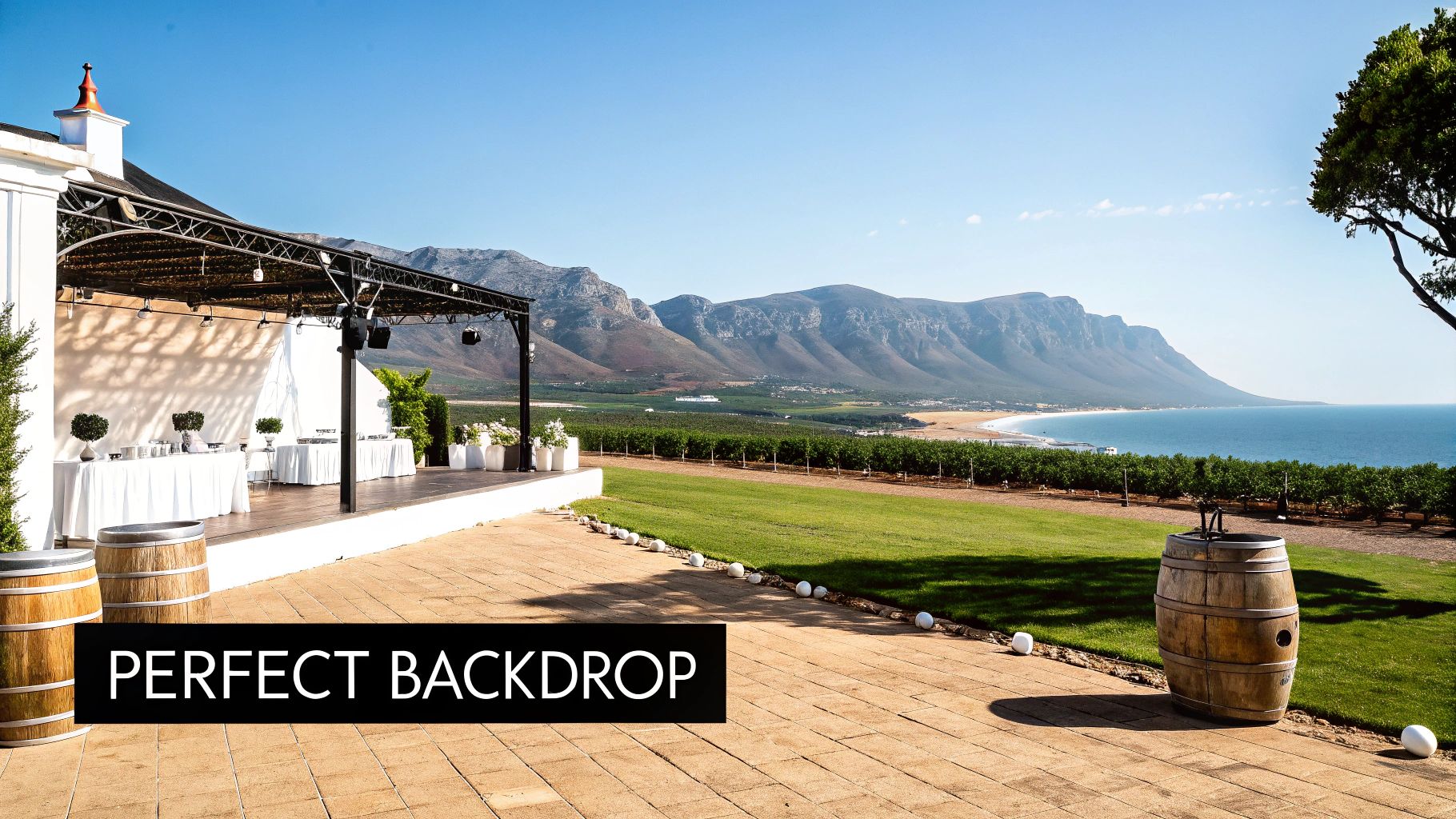 Elegant outdoor event space with white tables, a stage, wine barrels, and picturesque mountain and ocean views.