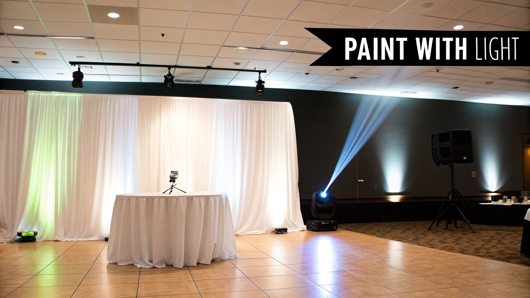 An indoor event space featuring white drapes, uplighting, a spotlight, and sound equipment.