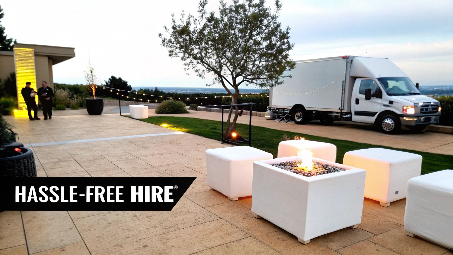 Outdoor patio at dusk with glowing fire pits, white lounge seating, string lights, and a white delivery truck.