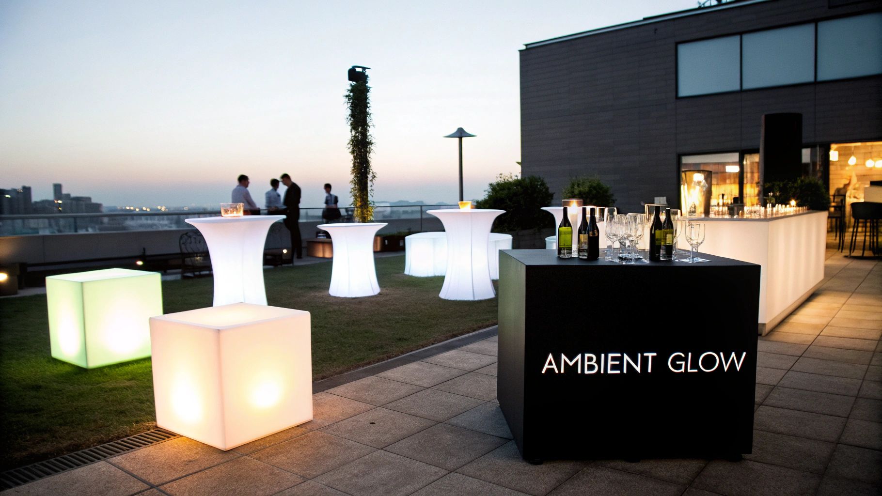 A modern rooftop lounge setup at dusk with illuminated tables, cubes, and a bar.
