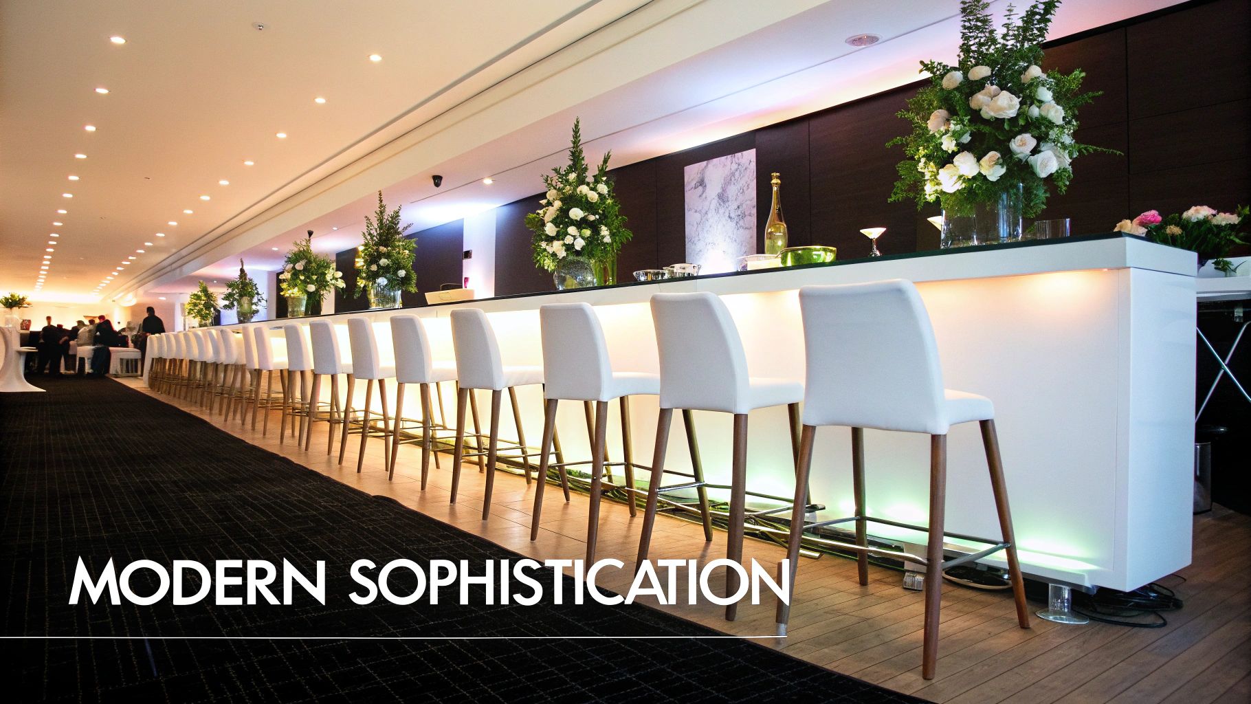 A long, modern bar counter illuminated with white lights, featuring numerous white bar stools and elegant floral arrangements.