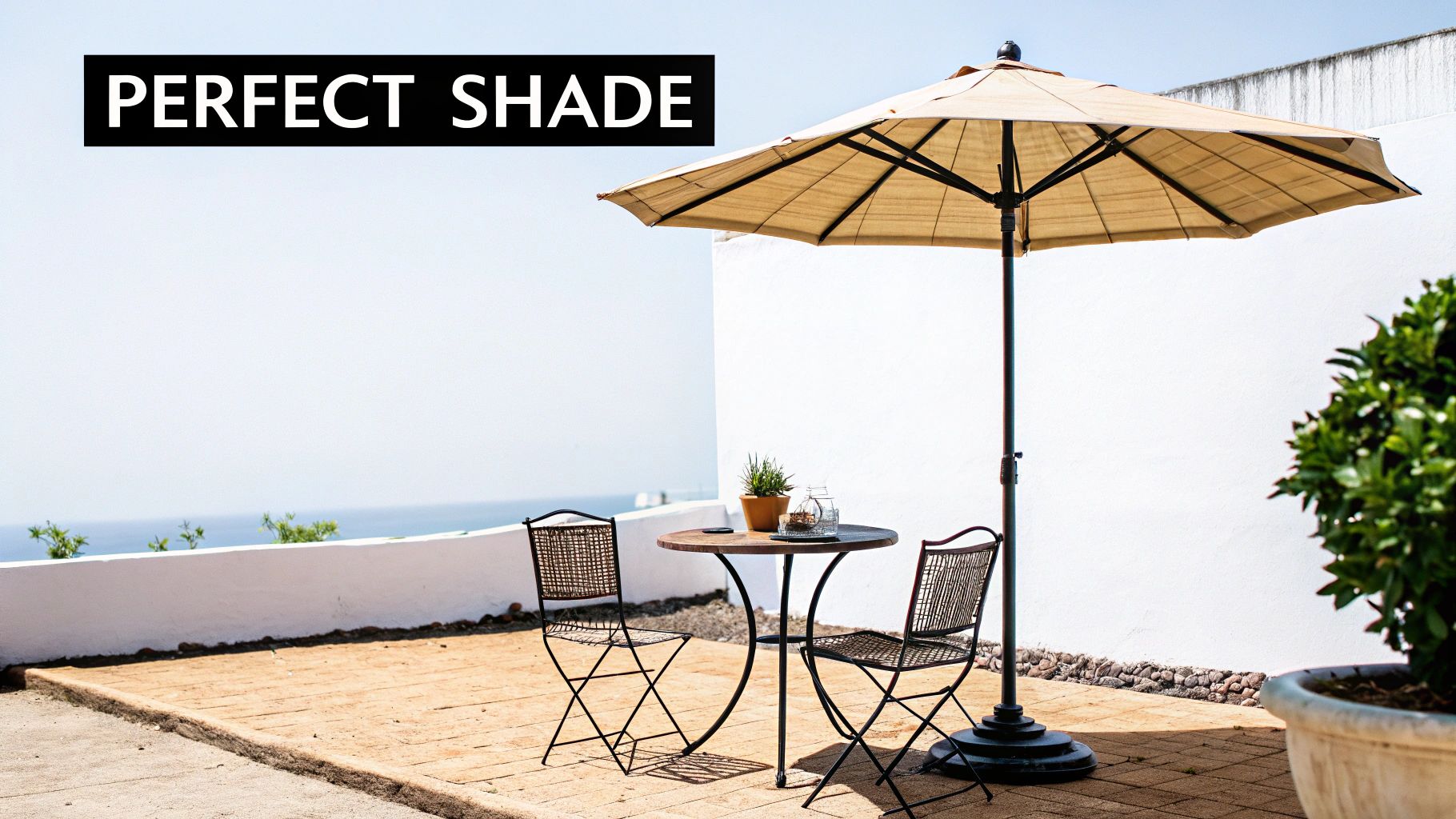 A beige outdoor umbrella shades a small round table and two chairs on a sunny patio overlooking the ocean.