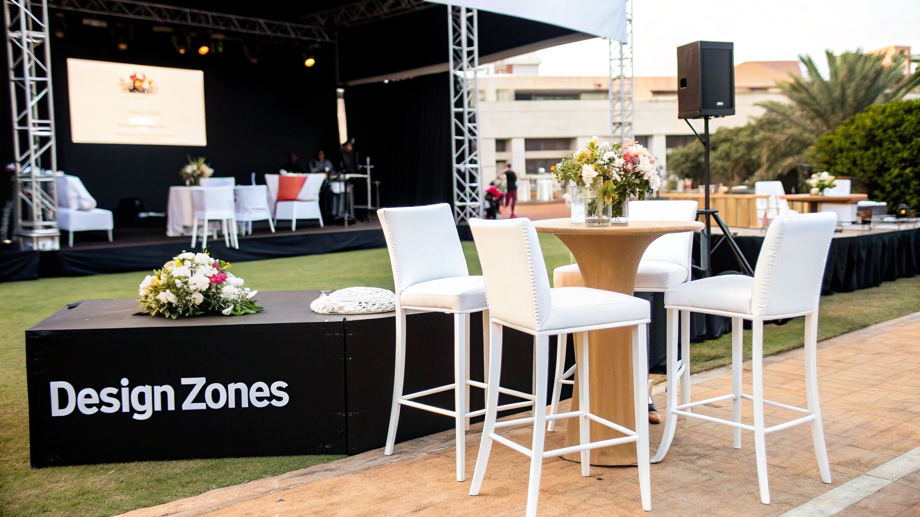Outdoor event setup with white bar chairs, a cocktail table, and a stage with a screen.