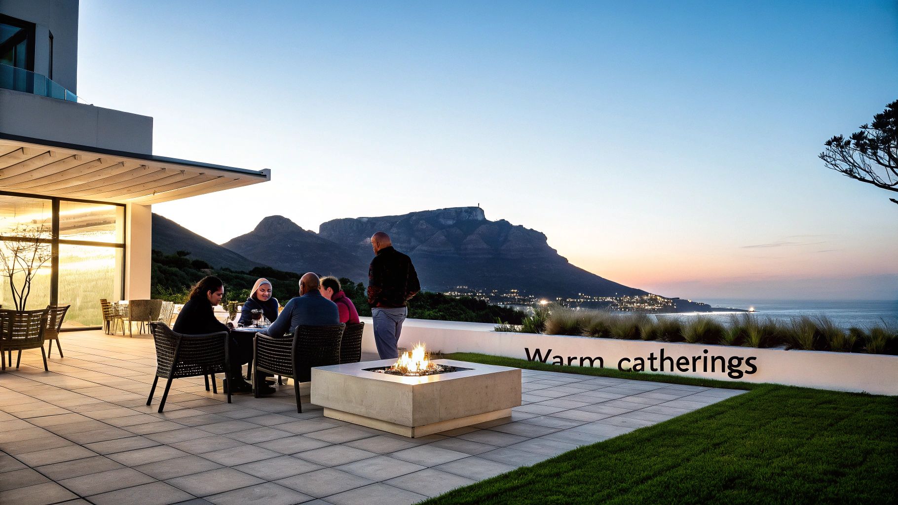 People gather on a patio around a glowing fire pit with a stunning mountain and ocean view at dusk.
