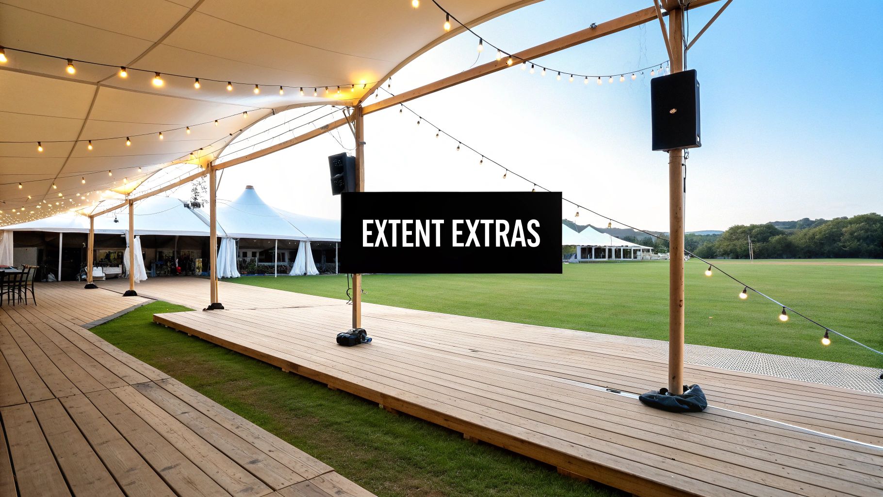 An outdoor event space with white stretch tents, wooden deck, string lights, and green field.