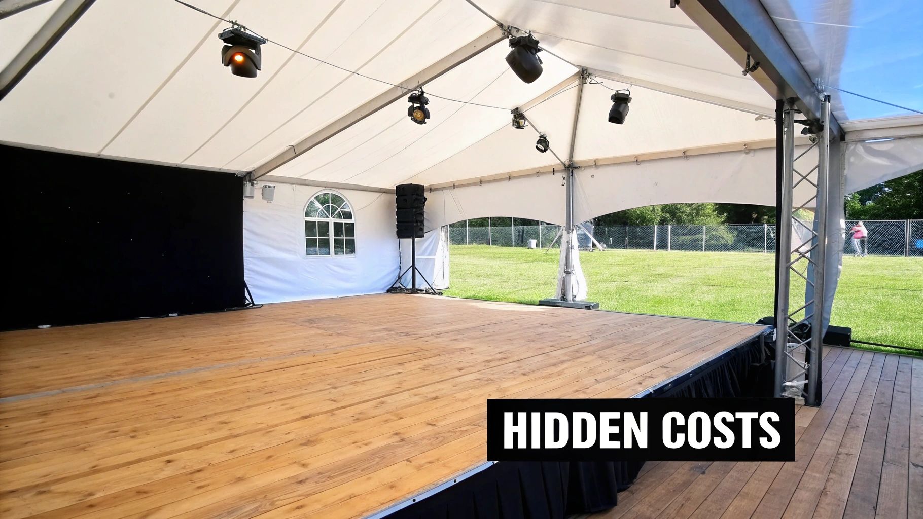An empty wooden stage is set up inside a large white event tent with lights and green grass outside.