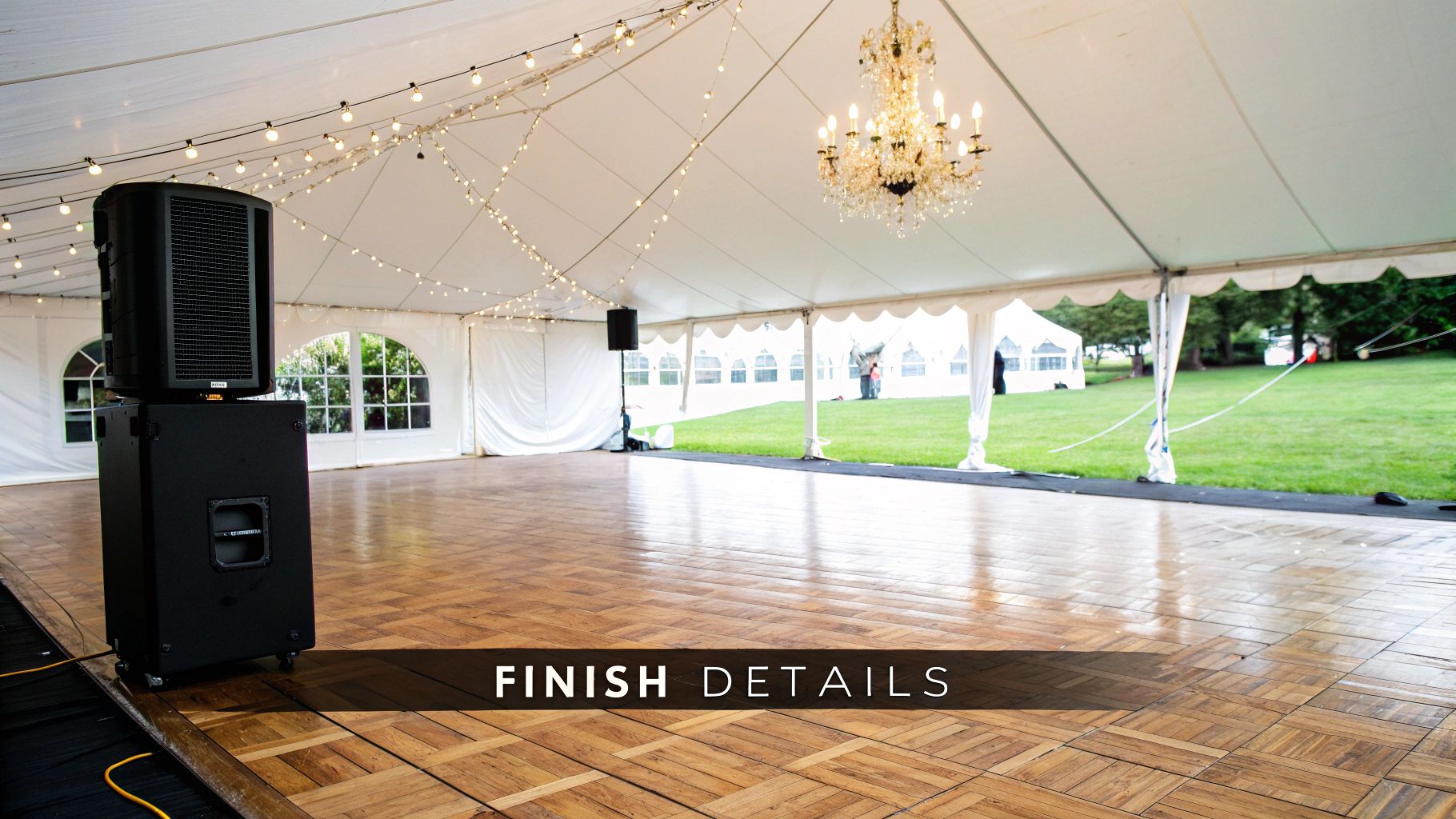Interior of a large white wedding tent with a polished wooden dance floor, elegant string lights, a chandelier, and speakers.