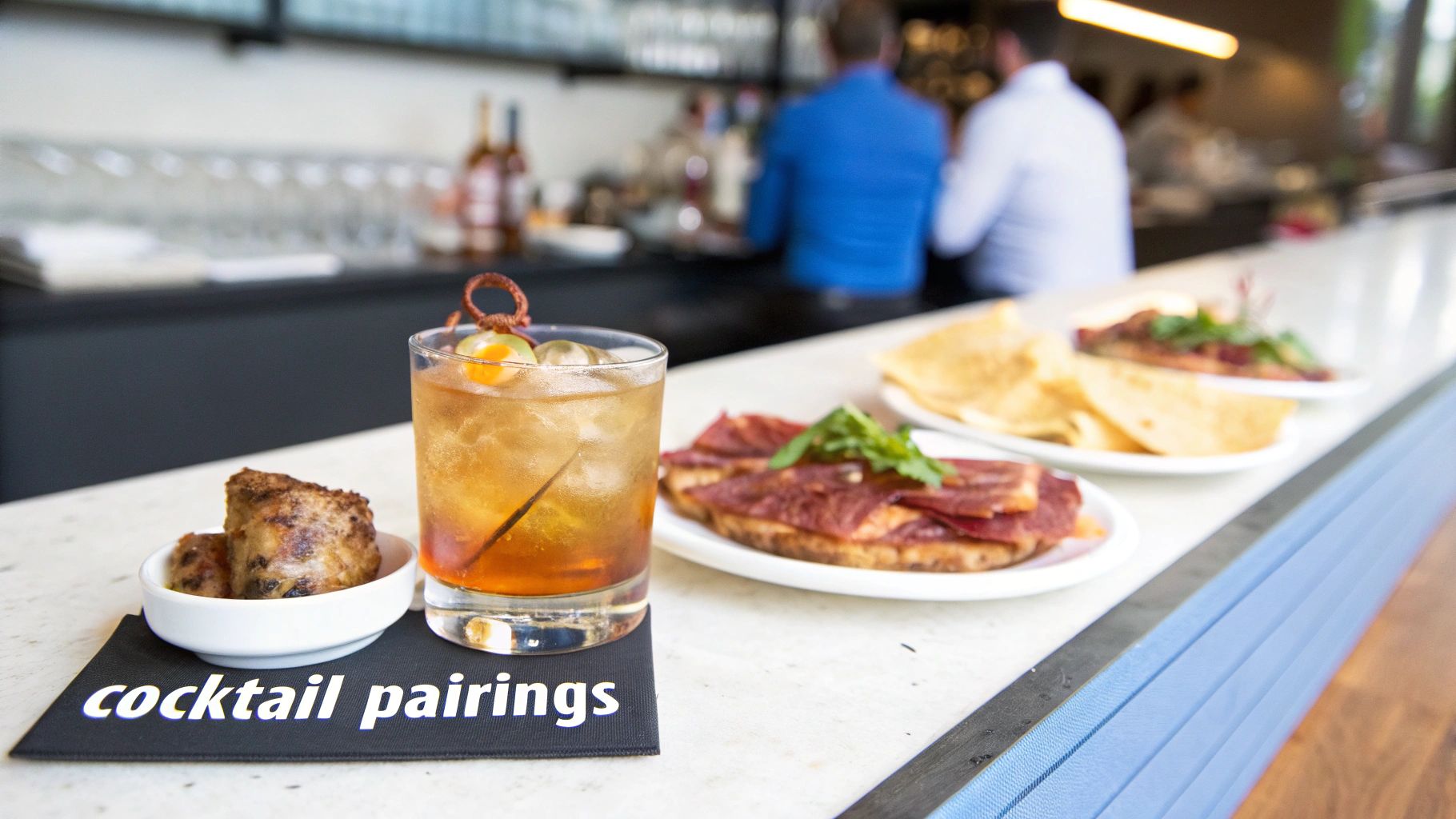 A bar counter displays various small plates of food and a refreshing cocktail with ice, indicating cocktail pairings.