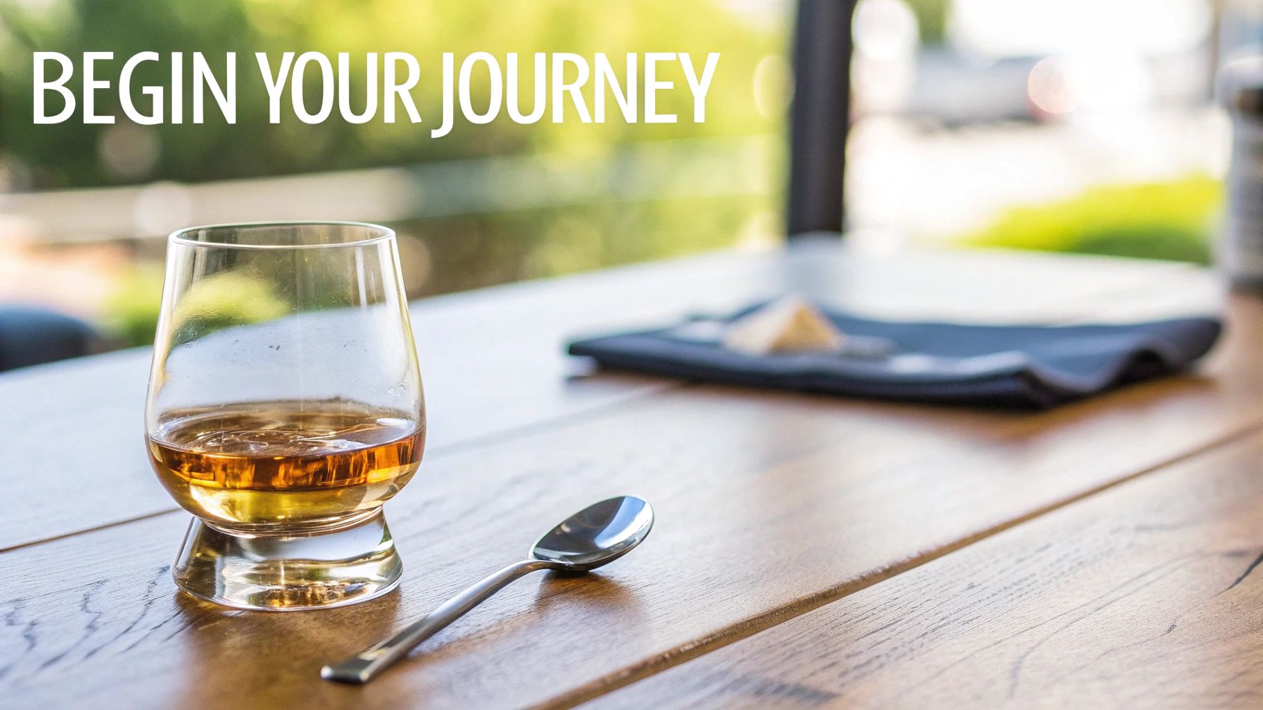 A glass of amber whiskey and a spoon on a wooden table with blurred food pairing in background.