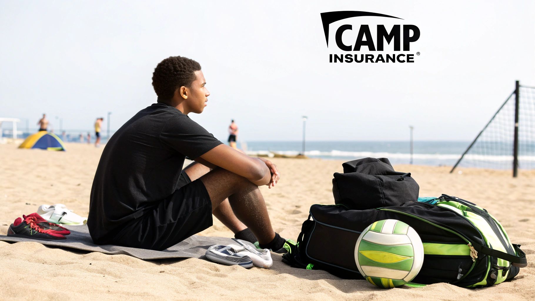 A young man sits on a sandy beach, looking at the ocean, with a volleyball and sports bag nearby.