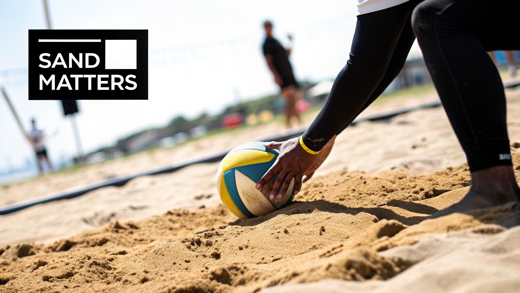 A person in black sportswear on a sandy beach volleyball court, holding a yellow and blue ball.